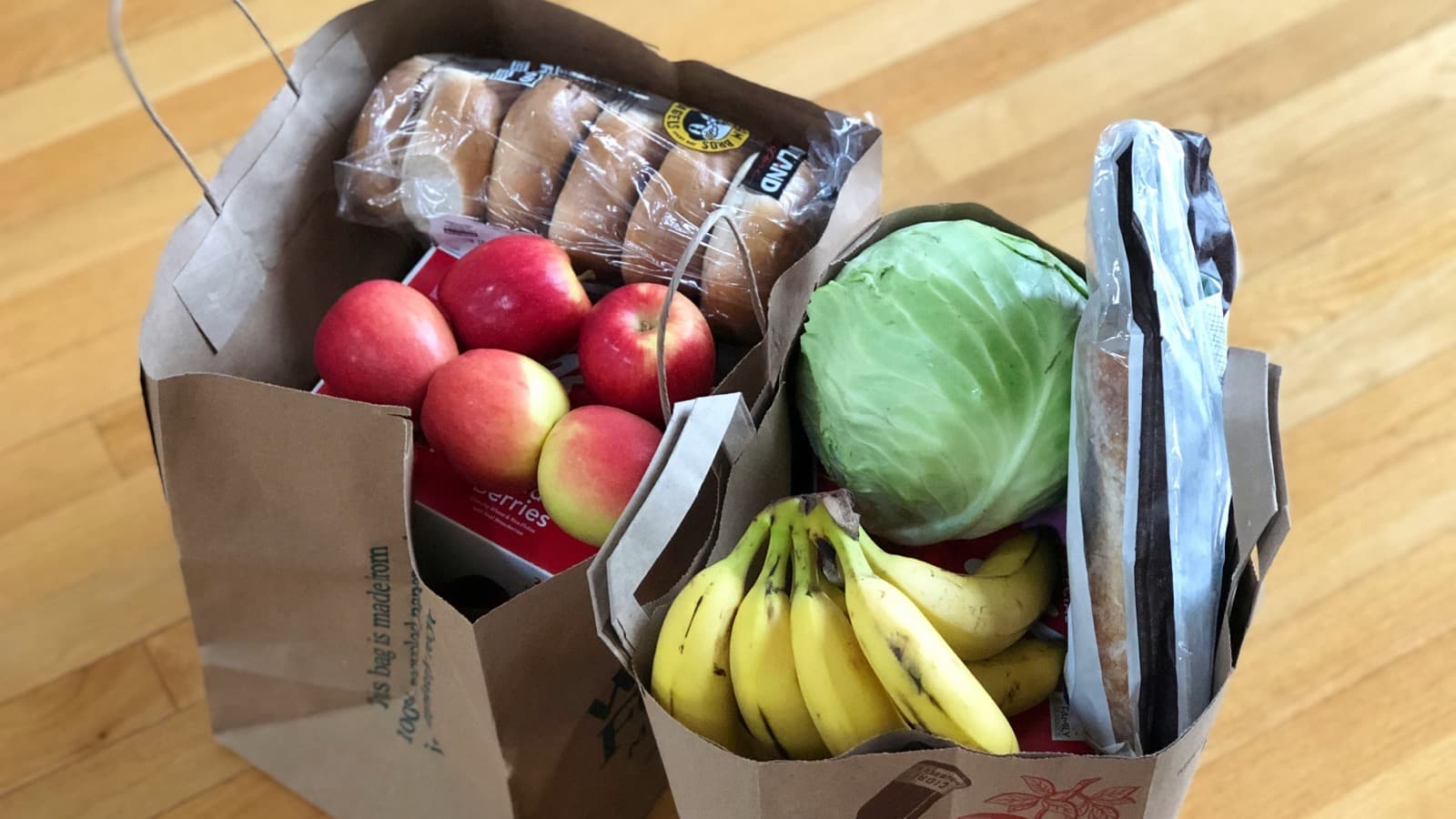 To papirposer med dagligvarer på et tregulv, med epler, bananer, hodekål og brød